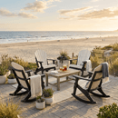 Four Kanyon Living Adirondack Rocking Chairs in Seashell & Black with blankets surround a wooden table with drinks on a patio overlooking the beach at sunset, with potted plants nearby. Made from durable poly lumber for all-weather use.
