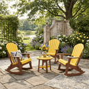 Three Kanyon Living Adirondack Rocking Chairs in yellow & cedar wood surround a small round table on a stone patio, decorated with flowers and cushions in a lush garden with trees, blooming bushes, and pond views.