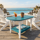 A Kanyon Living Poly Lumber Oval Coffee Table in Aruba Blue & White sits between two white Adirondack chairs on a wooden deck overlooking the ocean and palm trees, topped with magazines, a succulent plant, and iced tea on a sunny day.