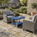 Two wicker armchairs with gray cushions rest on a stone patio, facing the Kanyon Living Poly Lumber Oval Coffee Table in Blue & Light Grey, topped with a mug and book. Lush gardens and a house appear in the background.