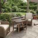 Wicker patio chairs with cream cushions surround the Kanyon Living Poly Lumber Oval Coffee Table in Brazilian Walnut & Weathered Wood on a stone patio, with plants, potted flowers, and a pergola in a lush garden. Gardening tools and a rolled mat are nearby.