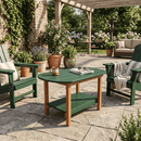 A sunlit patio features green armchairs and the Kanyon Living Poly Lumber Oval Coffee Table in dark green & cedar wood, handcrafted by Kanyon Living, set on a stone floor amid potted plants and outdoor furniture in a lush garden.