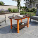 The Kanyon Living Poly Lumber Oval Coffee Table in Dark Wood & Orange holds two mugs, a magazine, and a phone. It's surrounded by gray patio chairs on a stone patio with garden plants and a house in the background.