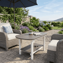 Outdoor patio with two wicker armchairs, a Kanyon Living Poly Lumber Oval Coffee Table Deluxe in Driftwood Grey & White topped with a potted plant, and a large umbrella. The space is framed by lush greenery, blooming flowers, and a scenic stone wall view.