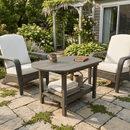 Two cushioned wicker chairs and a Kanyon Living Poly Lumber Oval Coffee Table Deluxe Driftwood Grey sit on a stone patio. The table holds mugs, a book, and folded blankets, with green plants and a white garden arbor in the background.