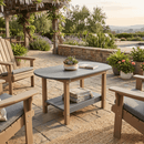 A cozy patio showcases wooden chairs and the Kanyon Living Poly Lumber Oval Coffee Table in Light Grey & Weathered Wood, topped with a potted plant and book. Stone flooring, greenery, and a pergola with hanging purple flowers enhance the inviting scene.