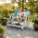 A smiling couple enjoys drinks on a sunny patio, seated on the Kanyon Living Poly Lumber 5′ Adirondack Glider Tall Version in Aruba Blue & White, with green plants and flowers around them and trees in the background.