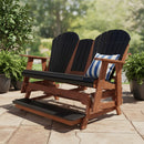 A Kanyon Living Poly Lumber 5′ Adirondack Glider Tall Version in Black and Deluxe Antique Mahogany sits on a patio amid potted plants, topped with a blue-and-white cushion, a drink on the armrest, and dappled sunlight from leafy trees.