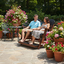 A man and woman smile on a wooden bench while the Kanyon Living Poly Lumber 5′ Adirondack Glider Tall Version in Black and Deluxe Antique Mahogany sits nearby, surrounded by colorful potted flowers on a stone patio in a lush garden.