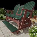 A Kanyon Living Poly Lumber 5′ Adirondack Glider Tall Version in Dark Green and Cherry Wood sits on a stone patio with potted white flowers; a round side table nearby holds a glass of iced tea.