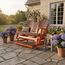 The Kanyon Living Poly Lumber 5′ Adirondack Glider Tall Version in Dark Wood and Orange offers inviting outdoor seating beside a patio table and potted hydrangeas, with a sunlit house and blooming garden softly glowing in the background.