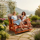 Two women relax on the Kanyon Living Poly Lumber 5′ Adirondack Glider Tall Version in Dark Wood and Orange, chatting amid potted flowers with drinks and a book. Mountains and water shine in the background, highlighting this ideal outdoor seating by Kanyon Living.