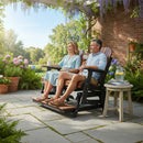 A smiling couple relaxes on the Kanyon Living Poly Lumber 5′ Adirondack Glider Tall Version Deluxe in Antique Mahogany and Black, enjoying drinks by blooming flowers and greenery in a sunny garden.