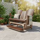 The Kanyon Living Poly Lumber 5′ Adirondack Glider Tall Version Deluxe in Brazilian Walnut and Weathered Wood sits on a sunny patio with potted plants, greenery, and a white lattice fence in the background.