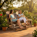 Two women relax and chat on the Kanyon Living Poly Lumber 5′ Adirondack Glider Tall Version Deluxe in Brazilian Walnut and Weathered Wood, enjoying drinks amid potted flowers on a sunny patio—a perfect scene with stylish outdoor furniture.