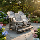 The Kanyon Living Poly Lumber 5′ Adirondack Glider Tall Version Deluxe Driftwood Grey with cushions and a throw blanket rests on a stone patio among blooming hydrangeas, potted plants, and a fountain, framed by lush trees at golden hour.
