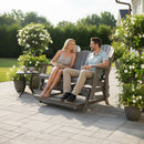 A man and woman sit smiling on the Kanyon Living Poly Lumber 5′ Adirondack Glider Tall Version Deluxe Driftwood Grey bench, surrounded by potted plants on a sunny patio, with a small table of coffee cups and lush greenery in the background.
