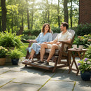 A smiling couple relaxes on the Kanyon Living Poly Lumber 5′ Adirondack Glider Tall Version in light grey and weathered wood, surrounded by lush plants and flowers, enjoying drinks on a small side table in their sunlit garden.