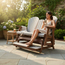 A woman in a light dress relaxes on the Kanyon Living Poly Lumber 5′ Adirondack Glider Tall Version (Light Grey and Weathered Wood), smiling with a mug on a sunny patio accented by greenery, flowers, and a small table.