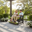 A smiling couple relaxes on a Kanyon Living Poly Lumber 5′ Adirondack Glider Tall Version in lime green and black, enjoying drinks and conversation on a sunny patio next to a small table with a pitcher and glasses.