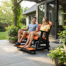 A smiling man and woman sit together on a Kanyon Living Poly Lumber 5′ Adirondack Glider Tall Version in orange and black, surrounded by potted plants on a stone patio outside a house with large windows.