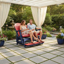 A smiling couple relaxes on the Kanyon Living Poly Lumber 5′ Adirondack Glider Tall Version in red and blue, under a pergola with white curtains, enjoying drinks on a sunny patio adorned with flowers and greenery.