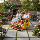 A smiling couple enjoys the Kanyon Living Poly Lumber 5′ Adirondack Glider Tall Version in red and yellow, surrounded by potted flowers. The man reads a book, the woman holds a drink, with a stone fireplace and green trees adding to the cozy scene.