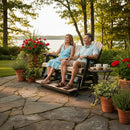 A smiling couple enjoys the Kanyon Living Poly Lumber 5′ Adirondack Glider Tall Version in Seashell and Black on a sunny patio, surrounded by flowers, greenery, and a lake view. Nearby sits a tray with mugs and a teapot.