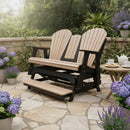 A Kanyon Living Poly Lumber 5′ Adirondack Glider Tall Version in Seashell and Black sits on a stone patio with potted purple hydrangeas and lavender. Nearby, a round table holds teacups and cookies amid lush, eco-friendly greenery.