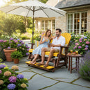 A smiling couple relaxes on the Kanyon Living Poly Lumber 5′ Adirondack Glider Tall Version in Yellow and Cedar Wood, enjoying drinks and snacks beneath a white umbrella in a hydrangea-filled garden with a stone house behind them.