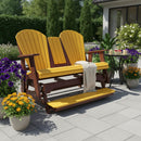 A Kanyon Living Poly Lumber 5′ Adirondack Glider Tall Version in Yellow and Cedar Wood sits on a patio, draped with a blanket over one armrest, surrounded by potted flowers, a small table with plants, and lush greenery in the background.