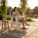 A woman in a light dress relaxes with a cup on the Kanyon Living Poly Lumber Adirondack Glider Chair Tall Deluxe in Driftwood Grey and White, placed on a rug-covered patio by a pond, with greenery and soft sunlight. A small table with a potted plant is beside her.