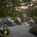 At dusk, three adults and a golden retriever relax in a lush garden under string lights, seated on Kanyon Living Poly Lumber Adirondack Glider Chair Tall Deluxe Driftwood Grey by a fire pit on the stone patio, surrounded by blooming hydrangeas.