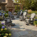 A stone patio with Kanyon Living Poly Lumber Adirondack Glider Chair Tall Deluxe Driftwood Grey arranged around a fire pit, surrounded by hydrangeas and greenery. A stone outdoor fireplace stands under a pergola.