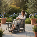 A woman relaxes in the Kanyon Living Poly Lumber Adirondack Glider Chair Tall Deluxe Driftwood Grey on a sunny patio, holding a mug. She's surrounded by potted plants, blooming flowers, string lights, and a small table nearby.