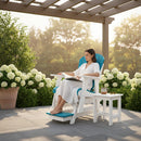 A woman in a white robe relaxes on a Kanyon Living Poly Lumber Adirondack Glider Chair Tall in Aruba Blue & White, reading a book under a pergola. Blooming white hydrangeas and a small white side table with drinks and glasses are nearby.
