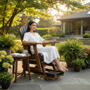 A woman in a white dress relaxes in the Kanyon Living Poly Lumber Adirondack Glider Chair Tall Black and Deluxe Antique Mahogany, surrounded by potted plants and flowers, with a book and cup on a nearby table amid stylish outdoor furniture.