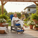 A woman in white smiles holding a book on the Kanyon Living Poly Lumber Adirondack Glider Chair Tall Blue and Light Grey, surrounded by potted plants on a deck under a pergola with string lights and eco-friendly outdoor furniture.