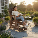 A woman relaxes outdoors on the Kanyon Living Poly Lumber Adirondack Glider Chair Tall Deluxe in Brazilian Walnut and Weathered Wood, reading a book among potted plants and poly lumber furniture as sunlight brightens the garden.