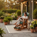 A woman relaxes, reading on the Kanyon Living Poly Lumber Adirondack Glider Chair Tall in Dark Green and Cedar Wood, surrounded by potted plants. Her mug sits on a side table as she enjoys her patio near large glass doors.