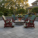 A stone patio with a round fire pit is encircled by four Kanyon Living Poly Lumber Adirondack Glider Chair Tall Dark Green and Cherry Wood pieces. A large potted plant with purple flowers sits behind the fire pit, framed by trees and a house.