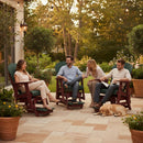 Four adults relax and socialize in a garden, seated on Kanyon Living Poly Lumber Adirondack Glider Chair Tall in Dark Green and Cherry Wood, surrounded by potted plants and string lights as a dog naps nearby, emphasizing the cozy outdoor vibe.