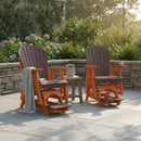Two Kanyon Living Poly Lumber Adirondack Glider Chairs in Tall Dark Wood and Orange with cushions and footrests are set on a stone patio by a low wall, with a small wooden table between them amid lush green plants and blooming flowers.