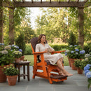 A woman relaxes barefoot with a mug on the Kanyon Living Poly Lumber Adirondack Glider Chair Tall Dark Wood and Orange under a pergola, surrounded by potted hydrangeas and greenery on a sunny patio.