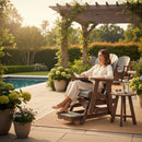 A woman relaxes on a Kanyon Living Poly Lumber Adirondack Glider Chair Tall in Light Grey and Weathered Wood by the garden pool, reading amid potted flowers and greenery, with a pergola and warm sunlight creating a peaceful scene.