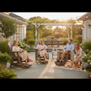Four adults relax on a sunny patio with flowers and string lights, while a golden retriever rests by the Kanyon Living Poly Lumber Adirondack Glider Chair in Tall Light Grey and Weathered Wood. An outdoor dining table stands in the background.