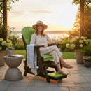 A woman in light clothing and a sunhat sits on the Kanyon Living Poly Lumber Adirondack Glider Chair Tall in Lime Green and Black, holding a mug, smiling at sunset. A blanket drapes over the eco-friendly chair’s arm amid patio plants and flowers.