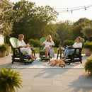 Three women relax and chat on Kanyon Living Poly Lumber Adirondack Glider Chair Tall Lime Green and Black outdoor furniture. A golden retriever rests nearby as string lights and potted plants complete the inviting stone patio scene.
