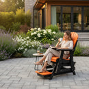 A woman relaxes on a Kanyon Living Poly Lumber Adirondack Glider Chair Tall Orange and Black with a mug and book. A side table holds her drink and snack, surrounded by flowers and a modern home for a cozy patio retreat.