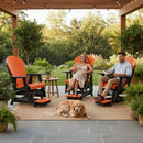 A woman and man relax on tall orange and black Kanyon Living Poly Lumber Adirondack Glider Chairs under a pergola with string lights, greenery, potted plants nearby, and a golden retriever resting on a rug at their feet.